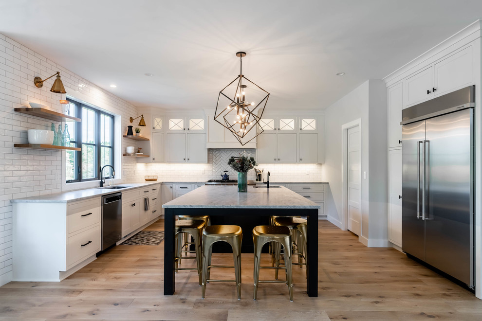This is an example of a country l-shaped kitchen in Portland Maine with an undermount sink, shaker cabinets, white cabinets, white splashback, subway tile splashback, stainless steel appliances, light hardwood floors, with island, beige floor and grey benchtop.
