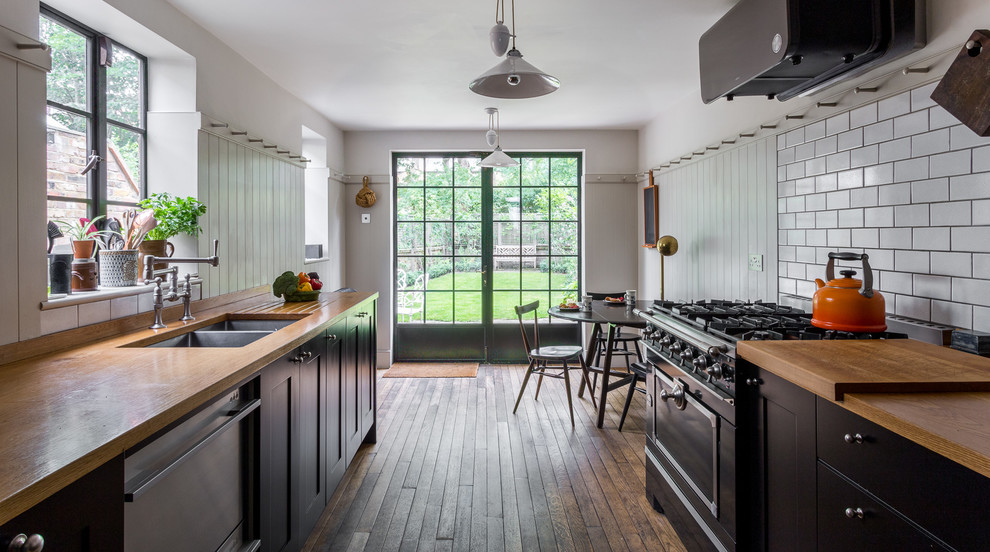 Mid-sized traditional galley eat-in kitchen in London with a double-bowl sink, black cabinets, wood benchtops, white splashback, subway tile splashback, black appliances, medium hardwood floors, shaker cabinets and no island.