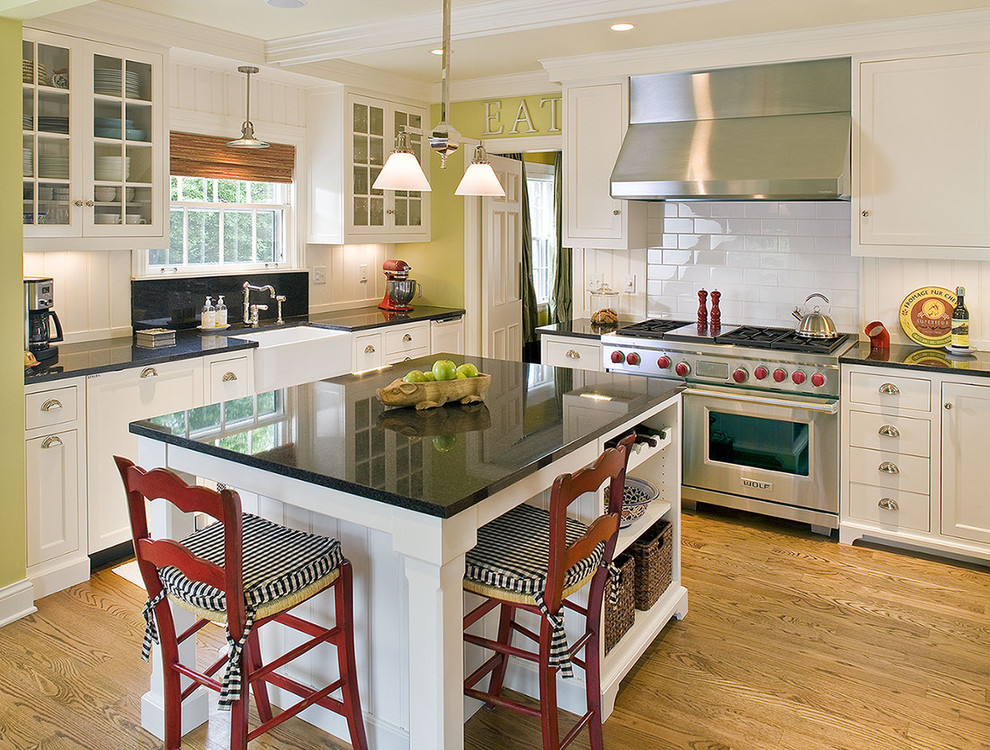 Mid-sized traditional u-shaped eat-in kitchen in Minneapolis with stainless steel appliances, a farmhouse sink, shaker cabinets, white cabinets, white splashback, subway tile splashback, light hardwood floors, with island and black benchtop.