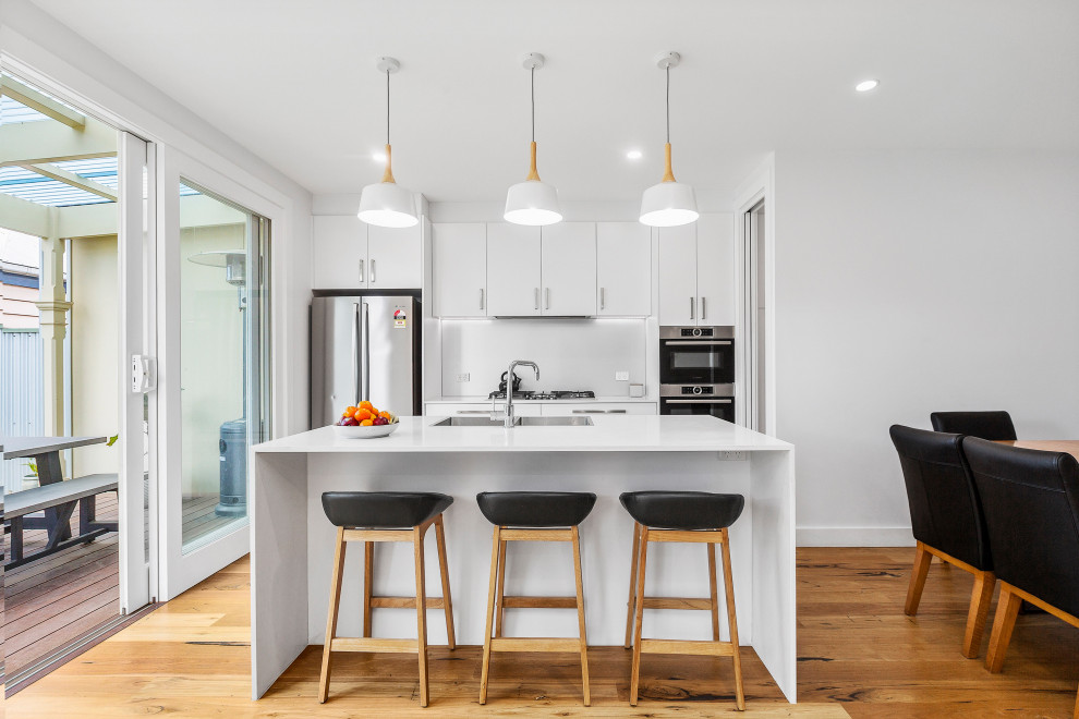 Mid-sized contemporary galley eat-in kitchen in Melbourne with a double-bowl sink, flat-panel cabinets, white cabinets, quartz benchtops, metallic splashback, glass sheet splashback, stainless steel appliances, medium hardwood floors, with island, brown floor and white benchtop.