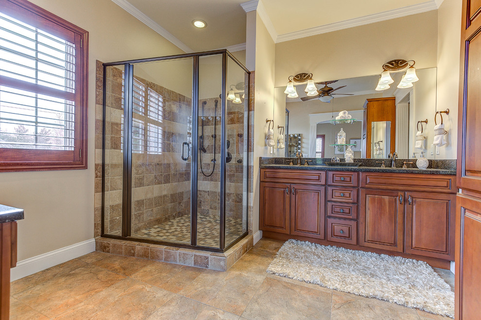 Photo of a large traditional master bathroom in Cincinnati with raised-panel cabinets, brown cabinets, a double shower, beige tile, travertine, travertine floors, an undermount sink, granite benchtops, beige floor, a hinged shower door, black benchtops and beige walls.