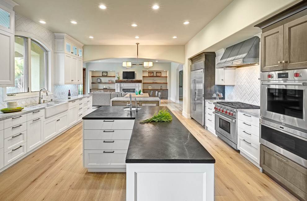 Large eclectic u-shaped open plan kitchen in Sacramento with a farmhouse sink, recessed-panel cabinets, white cabinets, marble benchtops, white splashback, porcelain splashback, stainless steel appliances, light hardwood floors and multiple islands.