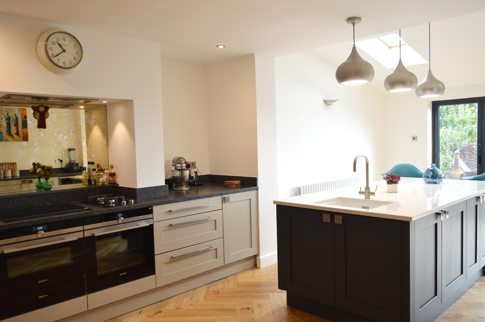 This is an example of a large contemporary l-shaped eat-in kitchen in Other with a drop-in sink, shaker cabinets, grey cabinets, granite benchtops, mirror splashback, black appliances, light hardwood floors and with island.