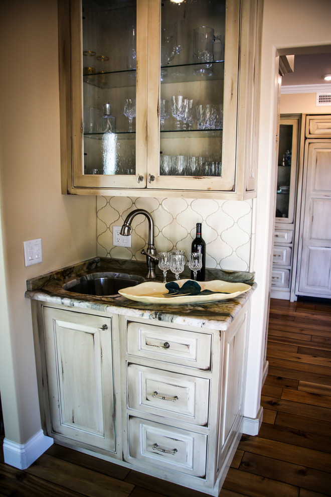Photo of a small country single-wall wet bar in Miami with a drop-in sink, raised-panel cabinets, distressed cabinets, marble benchtops, white splashback, plywood floors, brown floor and multi-coloured benchtop.