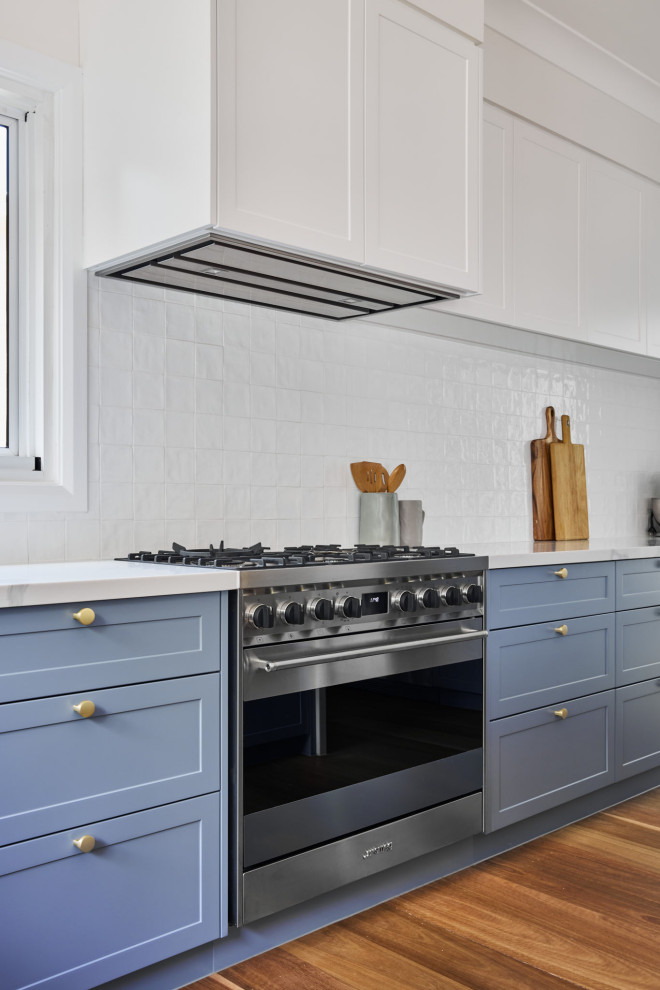 Large beach style u-shaped eat-in kitchen in Sydney with a farmhouse sink, shaker cabinets, blue cabinets, quartz benchtops, white splashback, engineered quartz splashback, stainless steel appliances, dark hardwood floors, with island, brown floor and white benchtop.