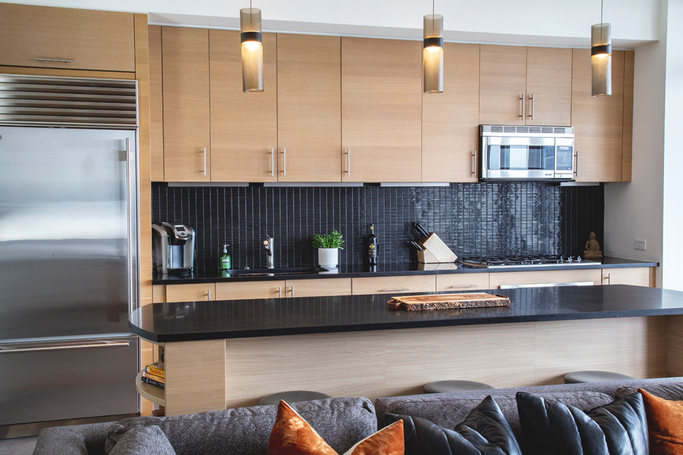 Modern single-wall open plan kitchen in New York with a drop-in sink, light wood cabinets, onyx benchtops, black splashback, mosaic tile splashback, stainless steel appliances, porcelain floors, with island, grey floor and black benchtop.