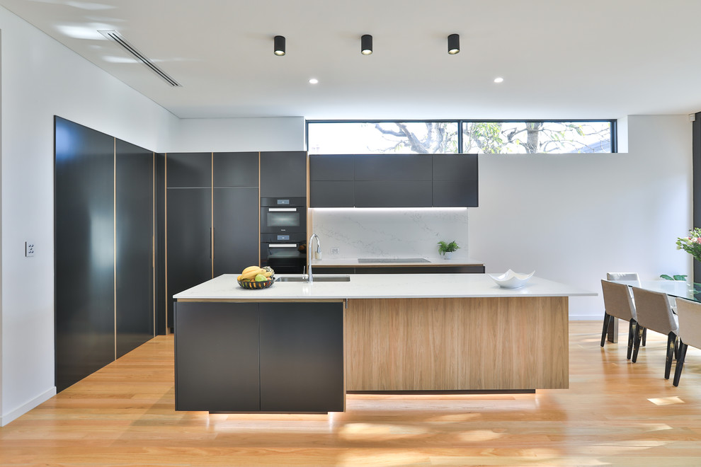 Inspiration for a large contemporary l-shaped eat-in kitchen in Sydney with an undermount sink, flat-panel cabinets, black cabinets, quartz benchtops, white splashback, stone slab splashback, black appliances, light hardwood floors, with island, white benchtop and brown floor.