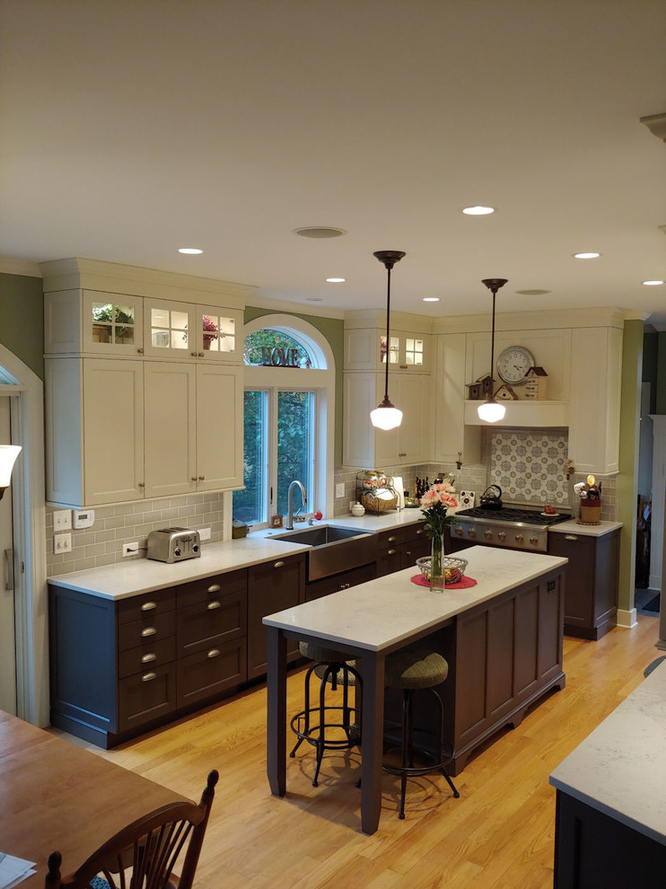 This is an example of a mid-sized traditional u-shaped eat-in kitchen in Boston with a farmhouse sink, recessed-panel cabinets, white cabinets, grey splashback, ceramic splashback, stainless steel appliances, medium hardwood floors, with island, brown floor and white benchtop.
