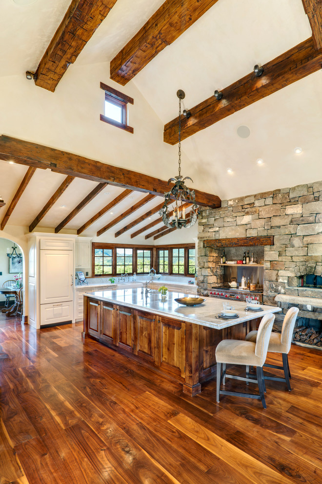 Large mountain style l-shaped medium tone wood floor open concept kitchen photo with a farmhouse sink, raised-panel cabinets, white cabinets, quartzite countertops, paneled appliances and an island