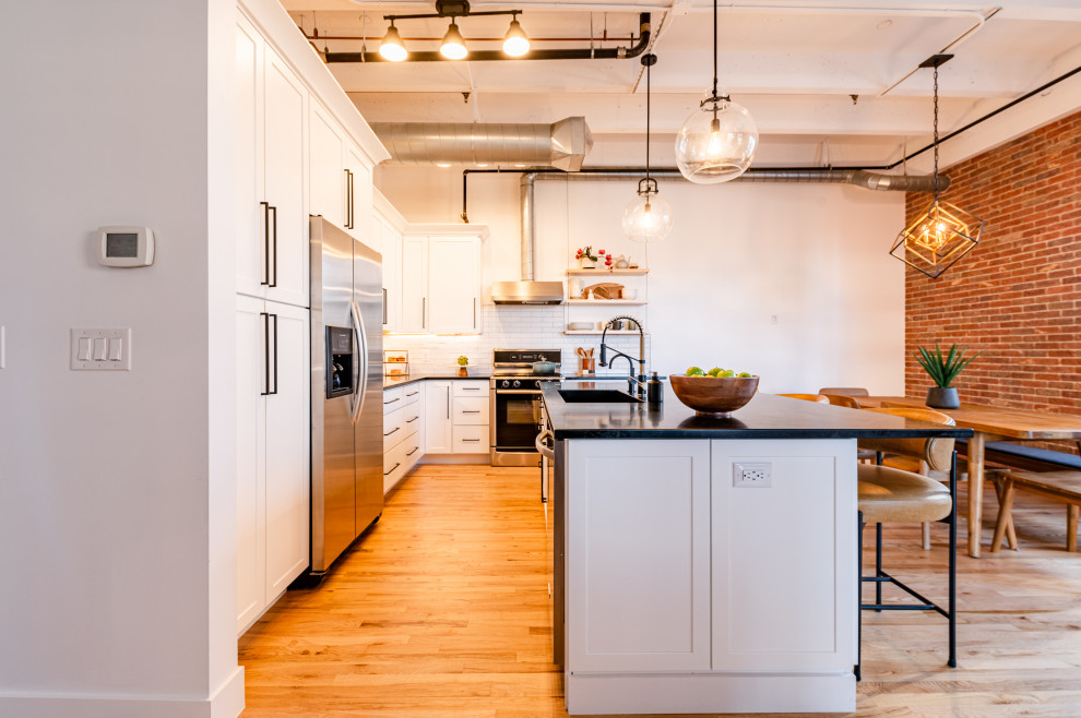 Design ideas for a large industrial l-shaped eat-in kitchen in Denver with an integrated sink, shaker cabinets, white cabinets, soapstone benchtops, white splashback, ceramic splashback, stainless steel appliances, light hardwood floors, with island, black benchtop and exposed beam.