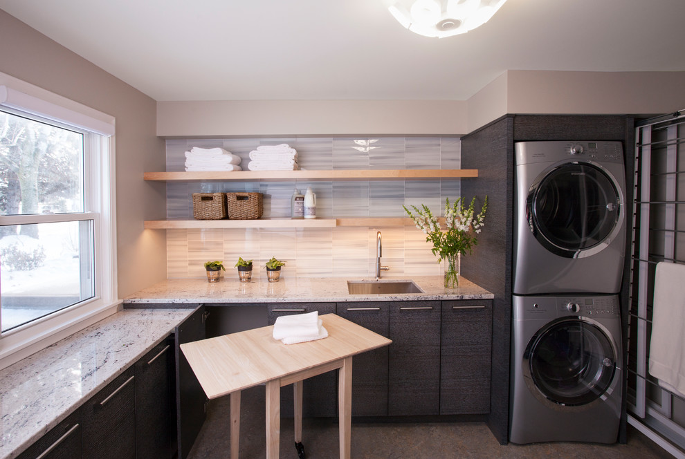 This is an example of a mid-sized modern l-shaped utility room in Minneapolis with an undermount sink, flat-panel cabinets, grey cabinets, granite benchtops, beige walls, laminate floors, a stacked washer and dryer and multi-coloured floor.