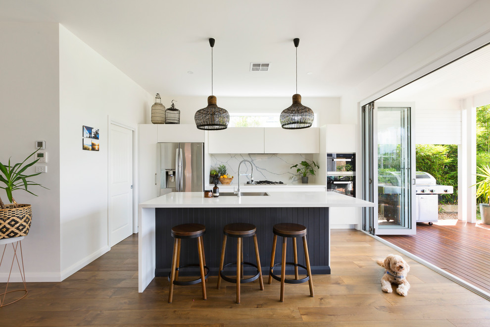 Beach style kitchen in Sydney with flat-panel cabinets, white cabinets, white splashback, stainless steel appliances, medium hardwood floors and with island.
