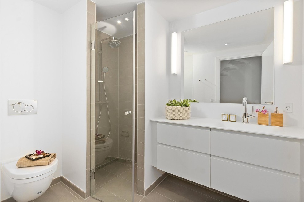 This is an example of a mid-sized scandinavian bathroom in Copenhagen with flat-panel cabinets, white cabinets, a wall-mount toilet, quartzite benchtops, a hinged shower door, a corner shower, beige tile, white walls, an integrated sink and white benchtops.