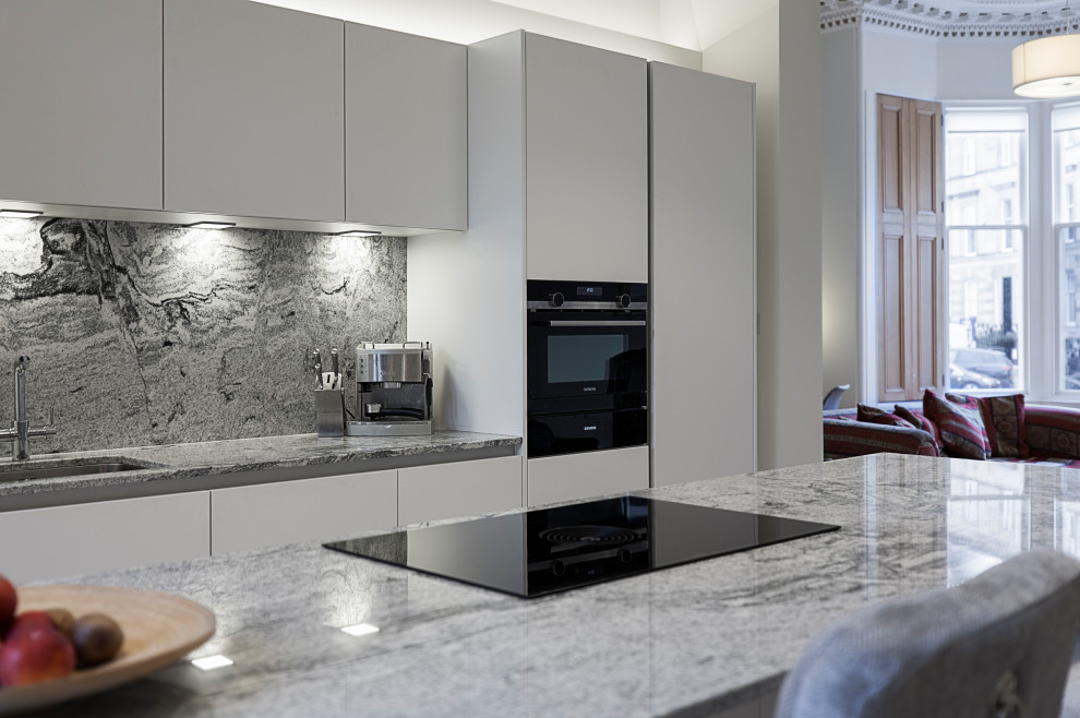 Photo of a large contemporary galley open plan kitchen in Glasgow with a single-bowl sink, flat-panel cabinets, white cabinets, granite benchtops, multi-coloured splashback, granite splashback, black appliances, medium hardwood floors, a peninsula and multi-coloured benchtop.