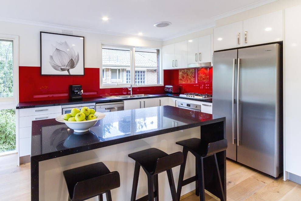 Design ideas for a mid-sized modern l-shaped eat-in kitchen in Melbourne with a drop-in sink, flat-panel cabinets, white cabinets, quartz benchtops, red splashback, glass sheet splashback, stainless steel appliances, light hardwood floors and with island.