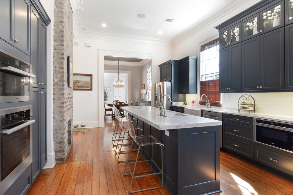 Large transitional eat-in kitchen in New Orleans with a farmhouse sink, recessed-panel cabinets, blue cabinets, white splashback, stainless steel appliances, light hardwood floors and with island.