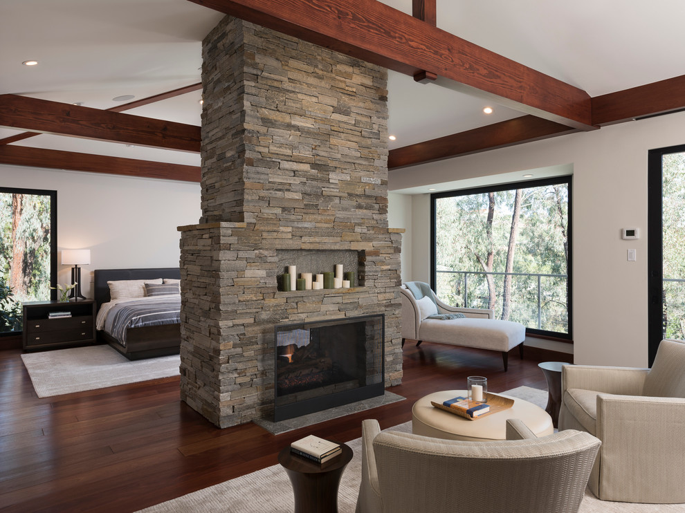 Photo of an expansive modern master bedroom in San Diego with white walls, medium hardwood floors, a two-sided fireplace, a stone fireplace surround and brown floor.