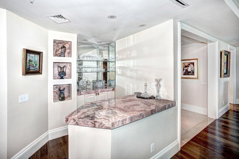 Inspiration for a mid-sized traditional single-wall wet bar in Miami with pink benchtop, no sink, raised-panel cabinets, white cabinets, granite benchtops, mirror splashback, dark hardwood floors and brown floor.