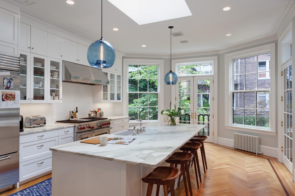 Design ideas for a transitional kitchen in New York with a farmhouse sink, glass-front cabinets, white cabinets, white splashback, stainless steel appliances, with island and medium hardwood floors.