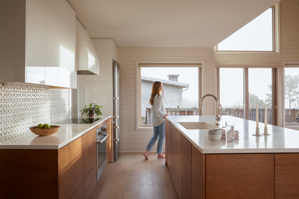 Mid-sized beach style galley light wood floor and beige floor open concept kitchen photo with a single-bowl sink, flat-panel cabinets, medium tone wood cabinets, quartz countertops, white backsplash, ceramic backsplash, stainless steel appliances, an island and white countertops