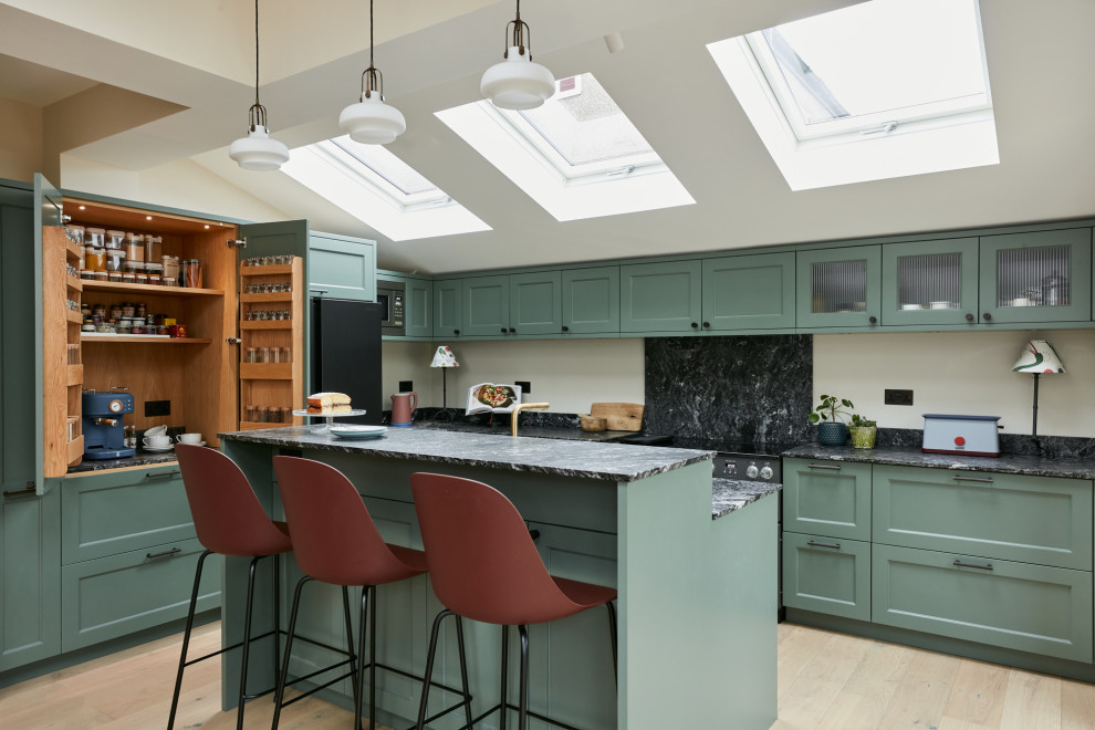 This is an example of a mid-sized transitional l-shaped open plan kitchen in London with a farmhouse sink, recessed-panel cabinets, green cabinets, granite benchtops, black splashback, granite splashback, light hardwood floors, with island, black benchtop and vaulted.