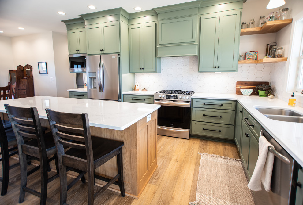 This is an example of a transitional l-shaped kitchen in Other with an undermount sink, shaker cabinets, green cabinets, quartz benchtops, white splashback, ceramic splashback, stainless steel appliances, laminate floors, brown floor and white benchtop.