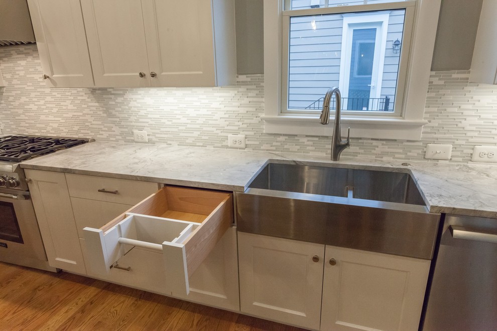 This is an example of a country l-shaped open plan kitchen in DC Metro with a farmhouse sink, shaker cabinets, white cabinets, quartzite benchtops, grey splashback, glass tile splashback, stainless steel appliances, light hardwood floors, with island, beige floor and grey benchtop.