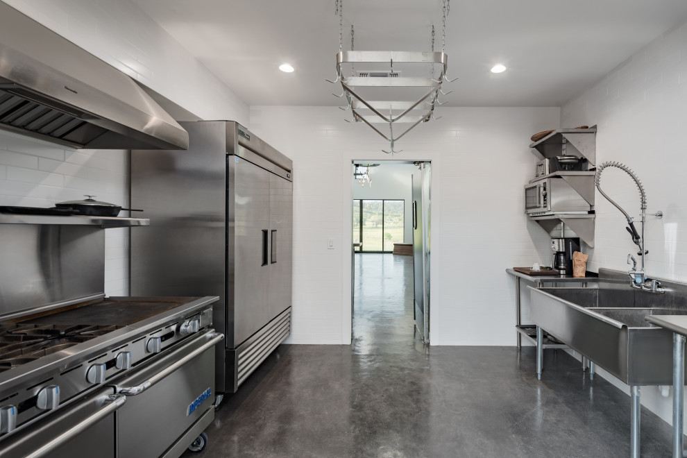 Photo of a large industrial separate kitchen in Austin with a triple-bowl sink, stainless steel benchtops, white splashback, ceramic splashback, stainless steel appliances, concrete floors and grey floor.
