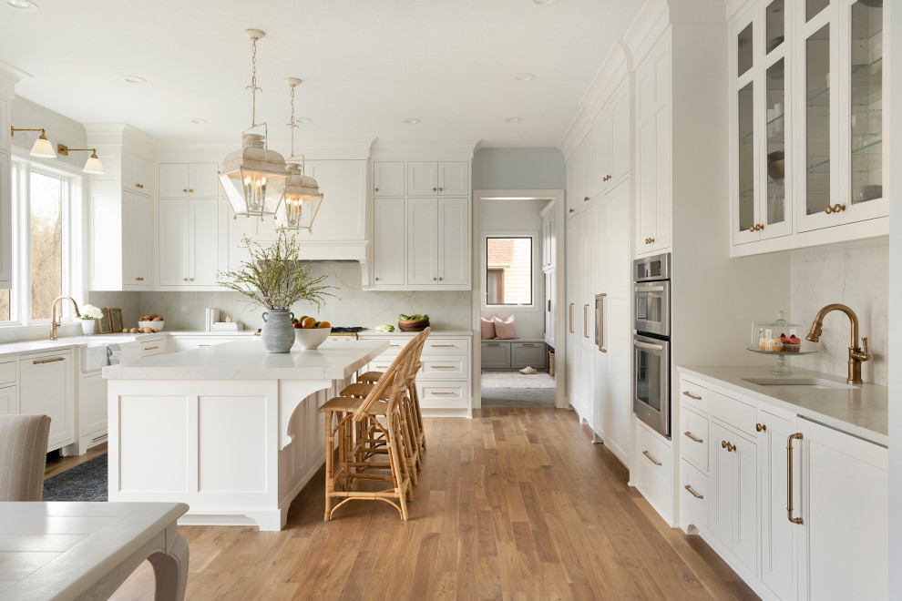 Inspiration for an expansive classic u-shaped kitchen/diner in Minneapolis with a belfast sink, recessed-panel cabinets, white cabinets, engineered stone countertops, white splashback, engineered quartz splashback, white appliances, light hardwood flooring, an island, brown floors and white worktops.