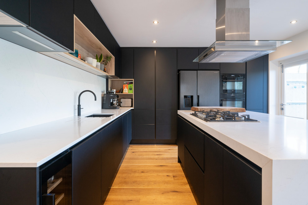 This is an example of a small contemporary l-shaped enclosed kitchen in Oxfordshire with a submerged sink, flat-panel cabinets, black cabinets, quartz worktops, white splashback, engineered quartz splashback, black appliances, light hardwood flooring, an island, brown floors, white worktops and a chimney breast.