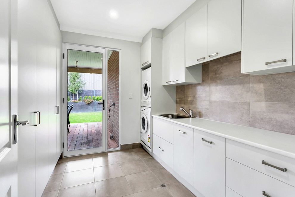 Photo of a large transitional galley utility room in Adelaide with a drop-in sink, flat-panel cabinets, grey cabinets, laminate benchtops, grey walls, porcelain floors, a stacked washer and dryer, grey floor and white benchtop.