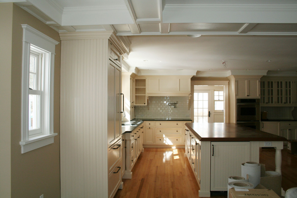 This is an example of a large country u-shaped eat-in kitchen in Boston with an undermount sink, recessed-panel cabinets, beige cabinets, soapstone benchtops, white splashback, subway tile splashback, panelled appliances, light hardwood floors, with island, beige floor and black benchtop.