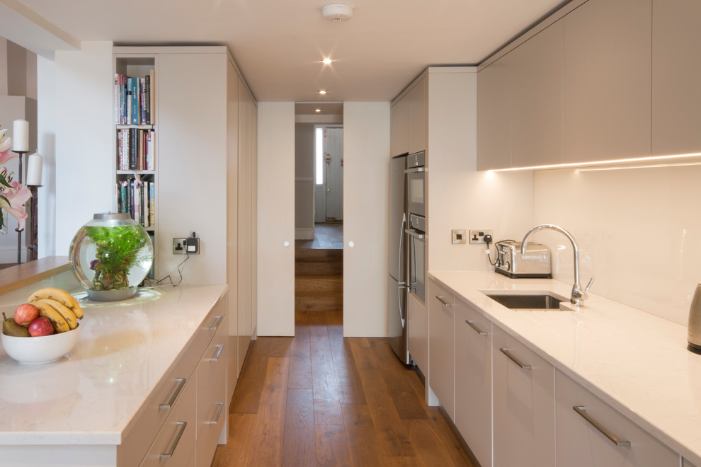 Design ideas for a mid-sized contemporary galley separate kitchen in Other with a drop-in sink, flat-panel cabinets, beige cabinets, quartz benchtops, white splashback, glass sheet splashback, stainless steel appliances and medium hardwood floors.