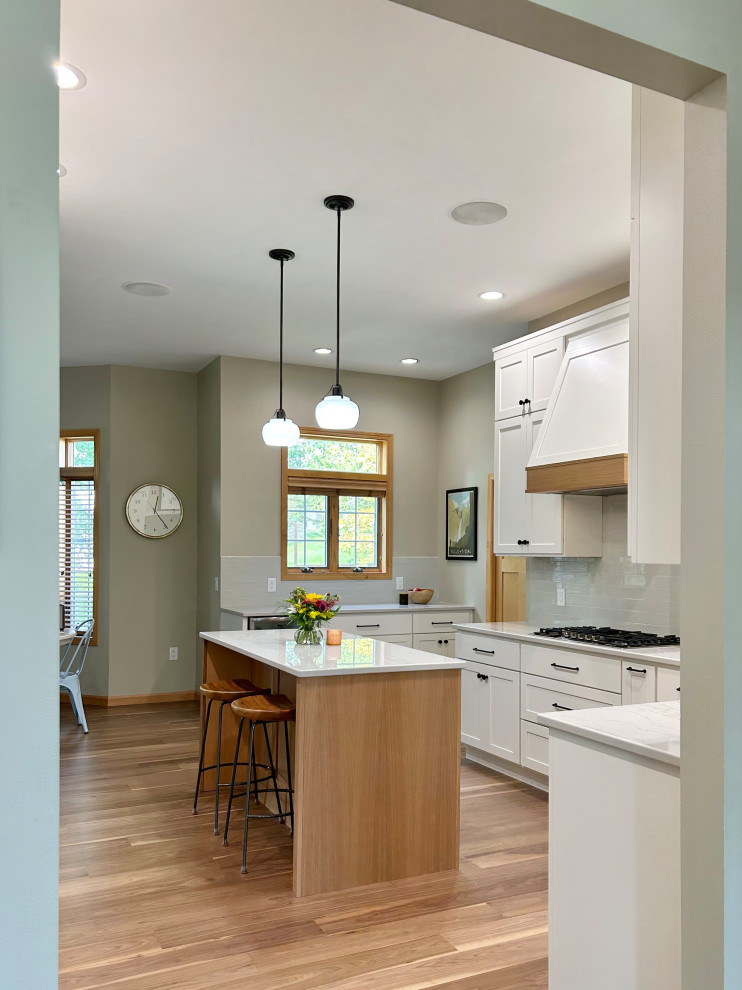 Example of a mid-sized transitional l-shaped medium tone wood floor and brown floor open concept kitchen design with an undermount sink, flat-panel cabinets, white cabinets, quartz countertops, gray backsplash, ceramic backsplash, stainless steel appliances, an island and white countertops