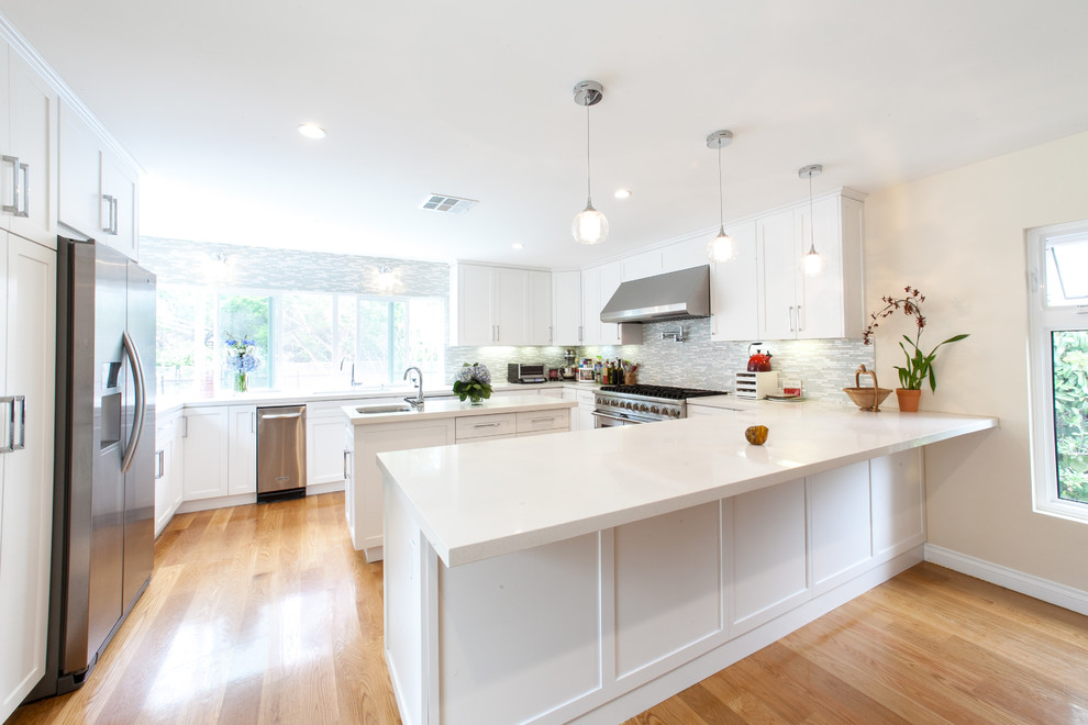 Design ideas for a large contemporary u-shaped open plan kitchen in Los Angeles with a double-bowl sink, shaker cabinets, white cabinets, quartz benchtops, grey splashback, mosaic tile splashback, stainless steel appliances, medium hardwood floors, multiple islands and brown floor.