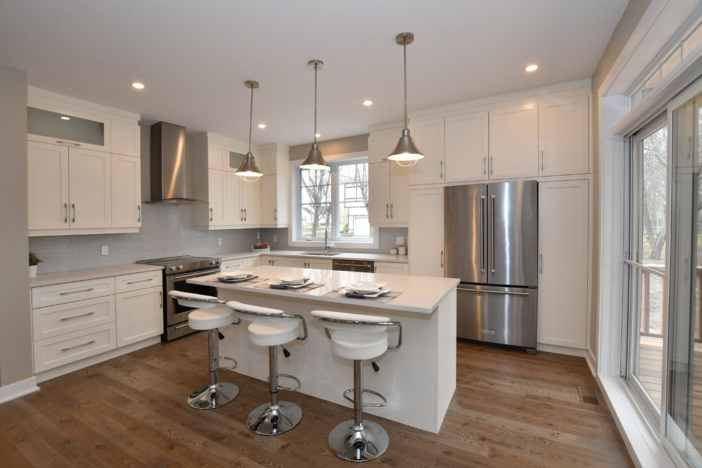 This is an example of a small transitional l-shaped open plan kitchen in Montreal with a double-bowl sink, shaker cabinets, white cabinets, quartz benchtops, multi-coloured splashback, glass tile splashback, stainless steel appliances, medium hardwood floors, with island and brown floor.