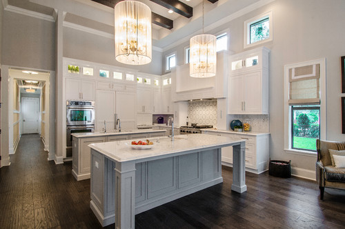 Spacious kitchen with ceiling-height white shaker RTA cabinets, glass transoms, twin marble-top islands, statement chandeliers, and dark hardwood floors.