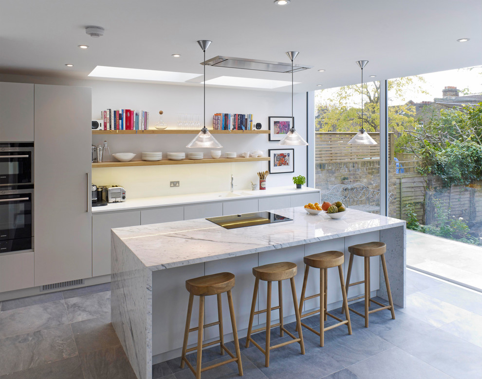 Large contemporary galley kitchen in London with an integrated sink, flat-panel cabinets, grey cabinets, marble worktops, white splashback, glass sheet splashback, ceramic flooring, an island and stainless steel appliances.