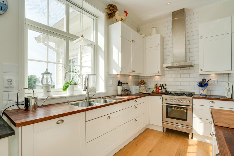 Inspiration for a mid-sized beach style l-shaped open plan kitchen in Gothenburg with a double-bowl sink, white cabinets, wood benchtops, white splashback, stainless steel appliances, light hardwood floors, no island, shaker cabinets and subway tile splashback.