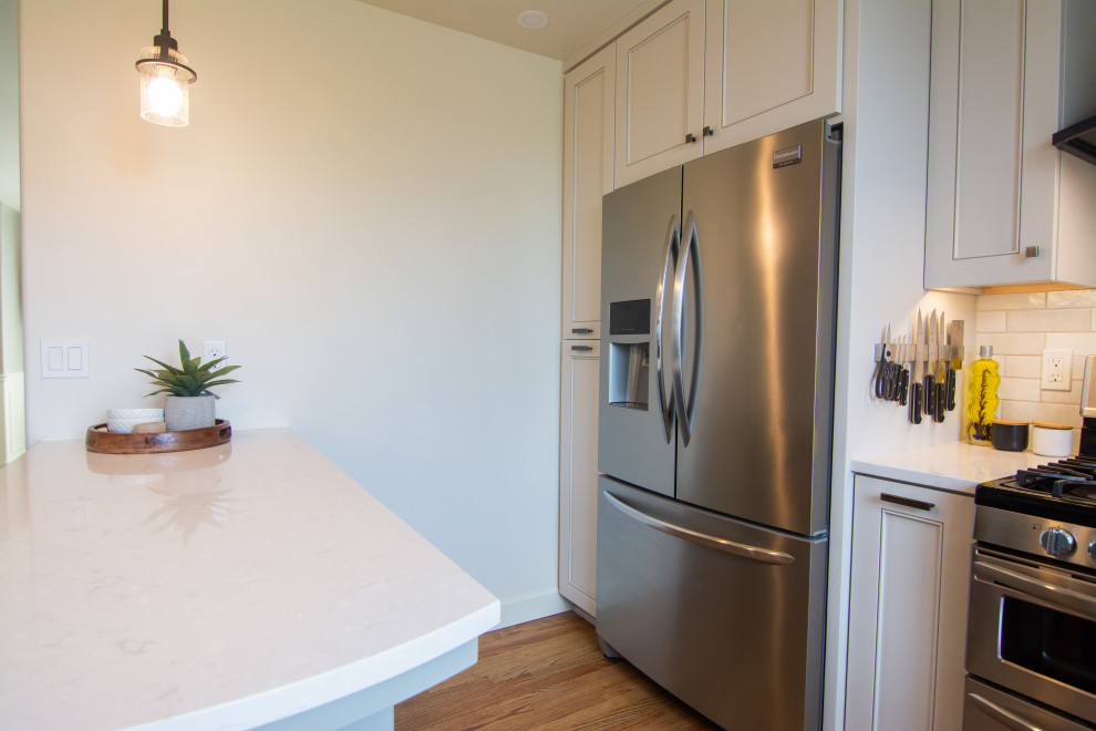 Example of a mid-sized transitional l-shaped medium tone wood floor and brown floor kitchen design with an undermount sink, recessed-panel cabinets, gray cabinets, quartz countertops, white backsplash, ceramic backsplash, stainless steel appliances, a peninsula and white countertops