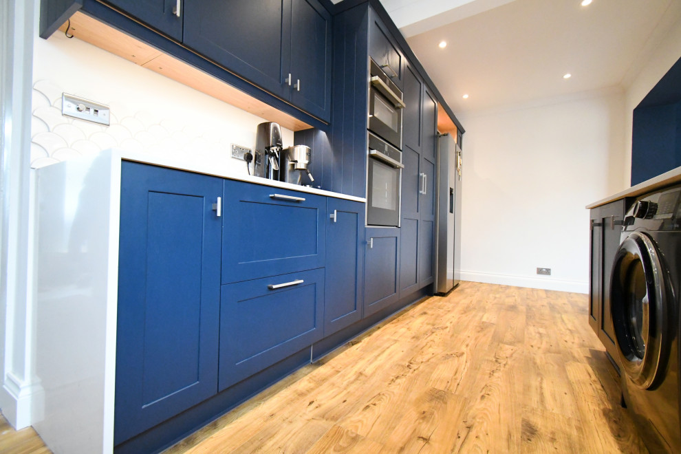 Photo of a mid-sized traditional galley eat-in kitchen in Essex with a farmhouse sink, shaker cabinets, blue cabinets, quartzite benchtops, white splashback, ceramic splashback, black appliances, laminate floors, no island, brown floor and white benchtop.