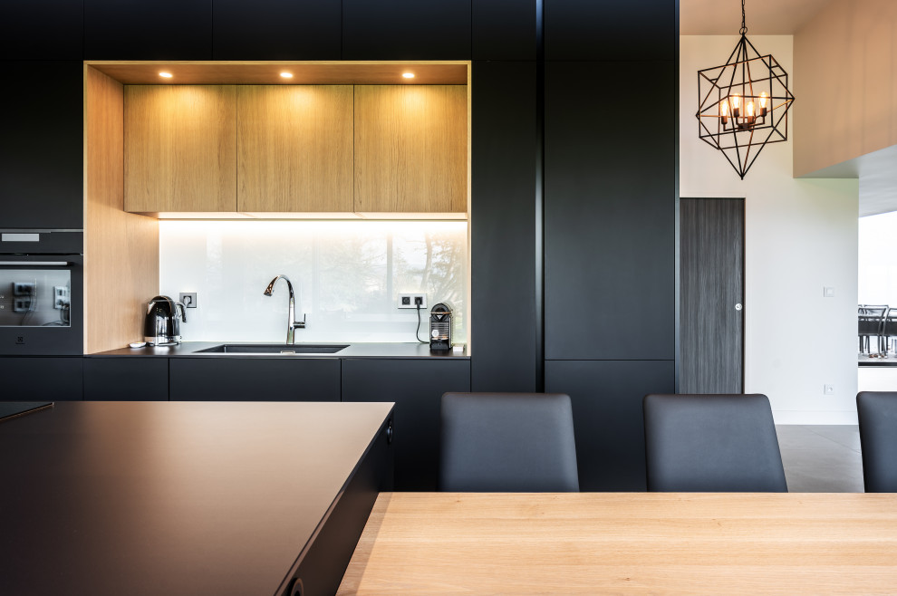 Large contemporary galley open plan kitchen in Lyon with an integrated sink, beaded inset cabinets, black cabinets, tile benchtops, white splashback, glass sheet splashback, black appliances, concrete floors, with island, black benchtop and coffered.
