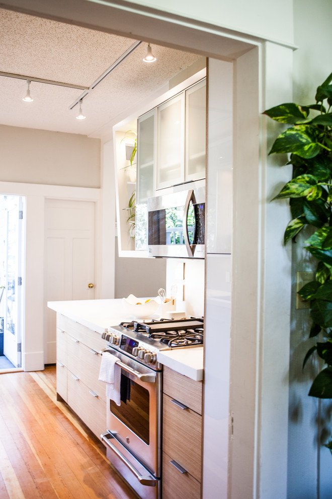 Medium sized modern galley kitchen/diner in Vancouver with a double-bowl sink, glass-front cabinets, light wood cabinets, composite countertops, metallic splashback, stainless steel appliances, light hardwood flooring and an island.
