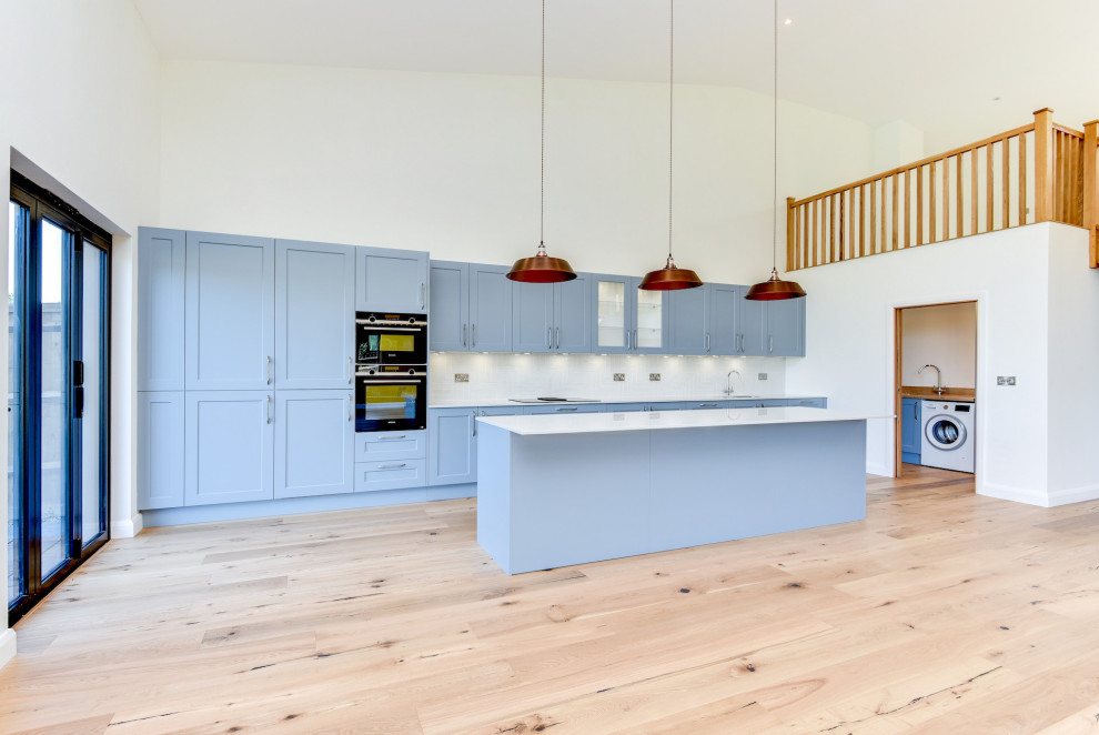 Design ideas for an expansive country galley open plan kitchen in Sussex with an undermount sink, shaker cabinets, blue cabinets, quartzite benchtops, white splashback, subway tile splashback, black appliances, light hardwood floors, with island, brown floor and white benchtop.