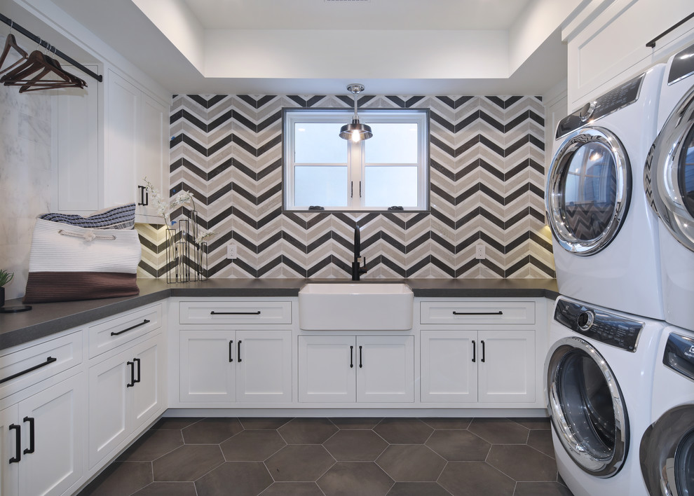 Inspiration for a transitional u-shaped dedicated laundry room in Orange County with a farmhouse sink, shaker cabinets, white cabinets, multi-coloured walls, a stacked washer and dryer, grey floor and grey benchtop.