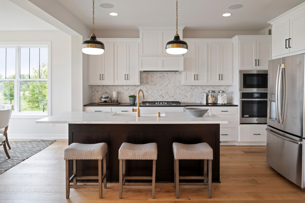 Design ideas for a large country l-shaped open plan kitchen in Minneapolis with a farmhouse sink, shaker cabinets, white cabinets, quartzite benchtops, white splashback, ceramic splashback, stainless steel appliances, with island, medium hardwood floors, brown floor and black benchtop.