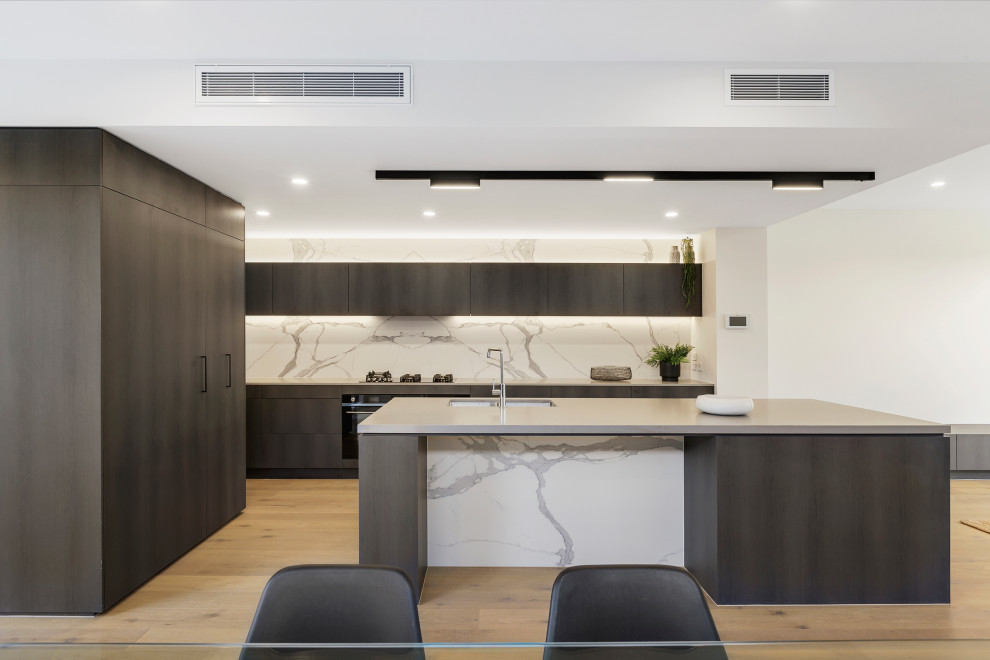 This is an example of a mid-sized contemporary galley open plan kitchen in Melbourne with an undermount sink, louvered cabinets, dark wood cabinets, concrete benchtops, multi-coloured splashback, porcelain splashback, black appliances, light hardwood floors, with island and grey benchtop.