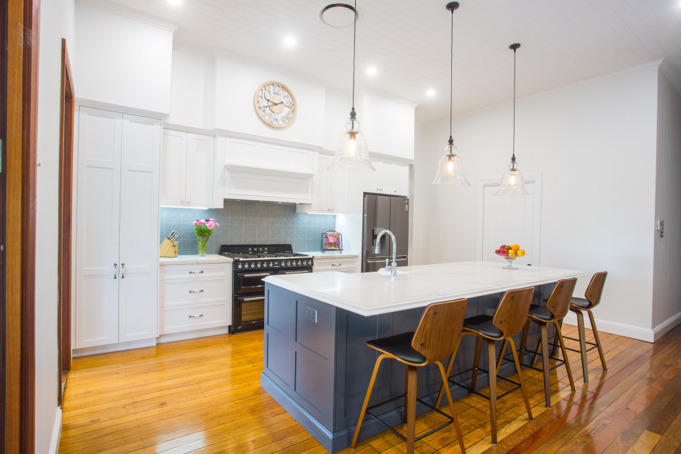 Photo of a large traditional galley open plan kitchen in Brisbane with a farmhouse sink, shaker cabinets, grey cabinets, quartz benchtops, grey splashback, subway tile splashback, black appliances, medium hardwood floors, with island, brown floor and white benchtop.