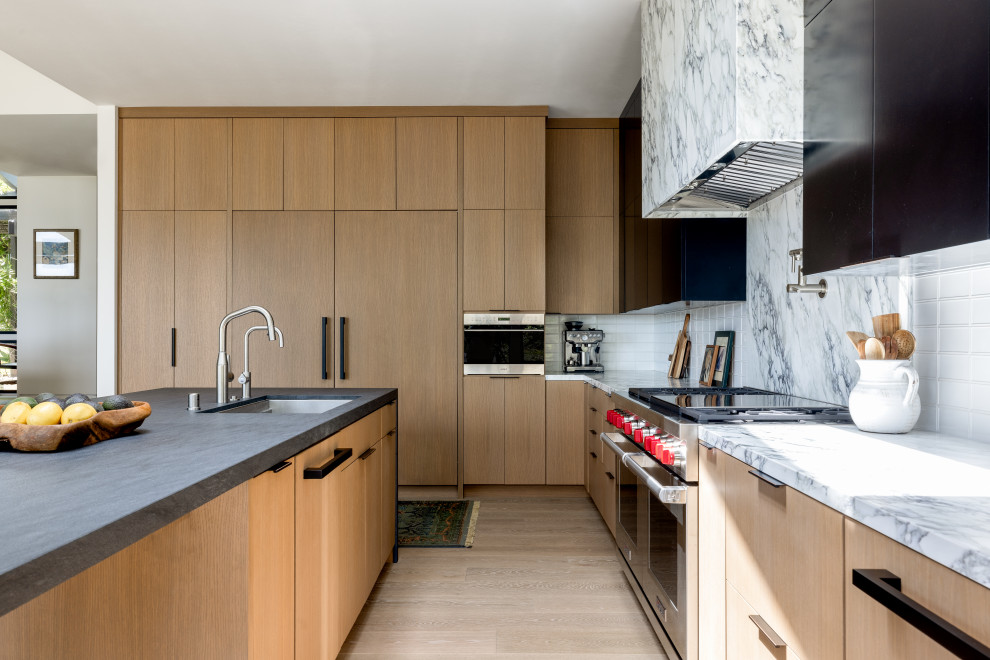 Example of a large minimalist l-shaped light wood floor and brown floor eat-in kitchen design in Santa Barbara with a drop-in sink, flat-panel cabinets, black cabinets, soapstone countertops, white backsplash, marble backsplash, stainless steel appliances, an island and black countertops