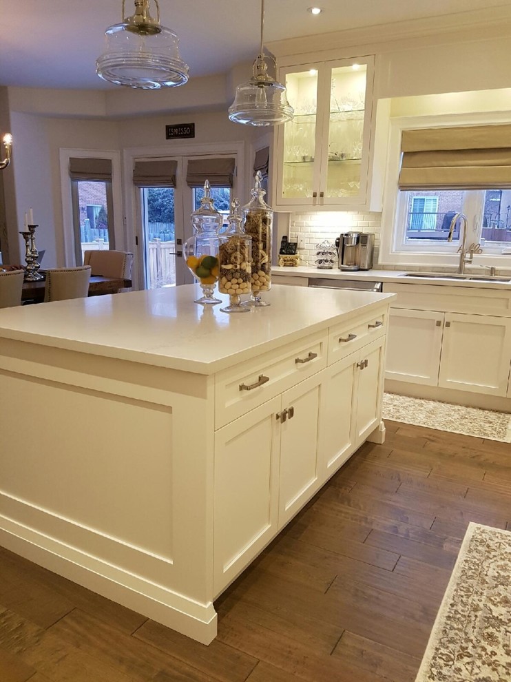 Open concept kitchen - mid-sized transitional l-shaped medium tone wood floor and brown floor open concept kitchen idea in Toronto with an undermount sink, beaded inset cabinets, white cabinets, solid surface countertops, white backsplash, brick backsplash, stainless steel appliances, an island and white countertops
